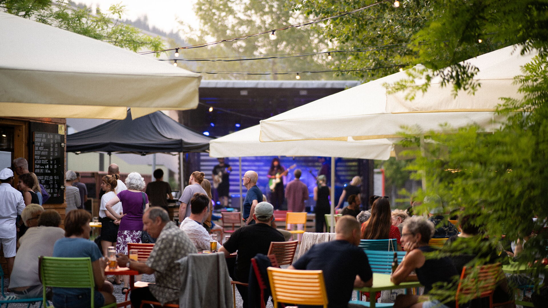 Menschen sitzen an bunten Tischen unter grossen Sonnenschirmen und schauen auf eine Bühne mit Musiker in einem Aussenbereich